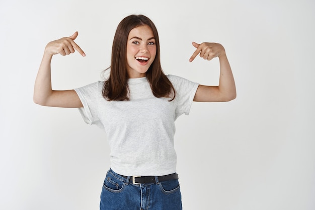 Jeune mannequin montrant une publicité, pointant du doigt le logo du t-shirt et souriant, debout sur un mur blanc.