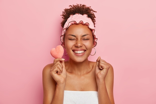 Jeune mannequin à la peau sombre et ravie, serre les poings de bonheur, tient une petite éponge douce en forme de cœur, ferme les yeux par plaisir, prête à prendre un bain, enveloppée dans une serviette porte un bandeau pour la douche