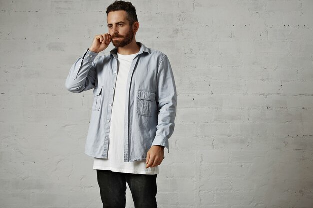 Jeune mannequin sérieux portant un t-shirt blanc, une chemise boutonnée en denim et un jean noir ajuste sa moustache isolé sur blanc