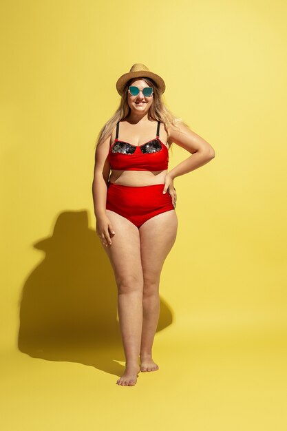 Jeune mannequin de taille plus caucasienne prépare des vacances sur un mur jaune. Femme en maillot de bain rouge, chapeau et lunettes de soleil.