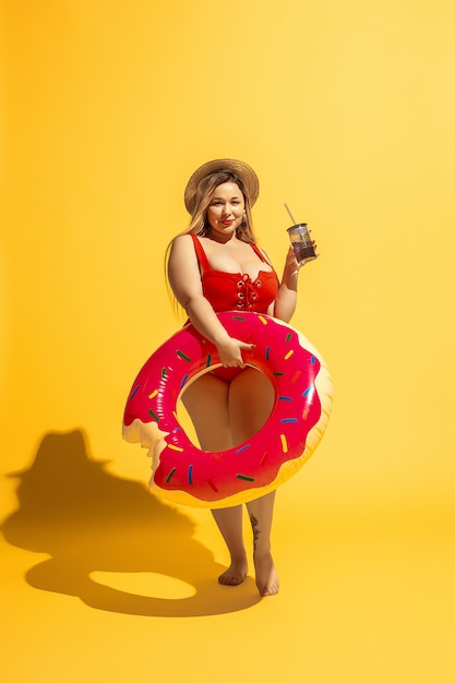 Jeune mannequin de taille plus caucasienne prépare des vacances sur un mur jaune. Femme en maillot de bain rouge et chapeau posant avec la natation.