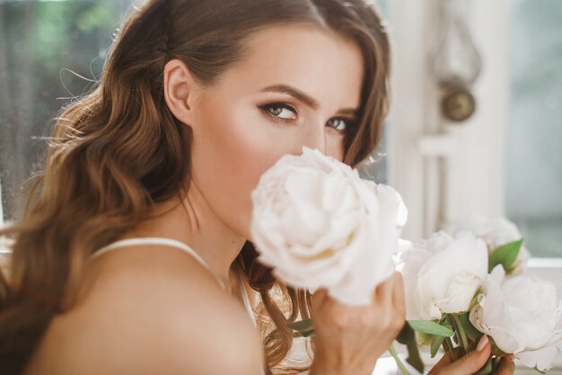 jeune mariée tient un bouquet de pivoines emplacement sur le rebord de la fenêtre dans le matin lumineux