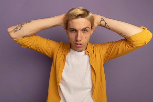 Jeune mec blond regardant droit devant portant un t-shirt jaune mettant les mains derrière la tête isolé sur violet