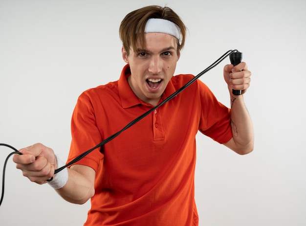 Jeune mec sportif en colère portant bandeau avec bracelet qui s'étend de la corde à sauter isolé sur un mur blanc