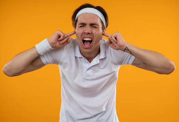 Jeune mec sportif en colère avec les yeux fermés portant bandeau et bracelet oreilles fermées isolé sur mur orange