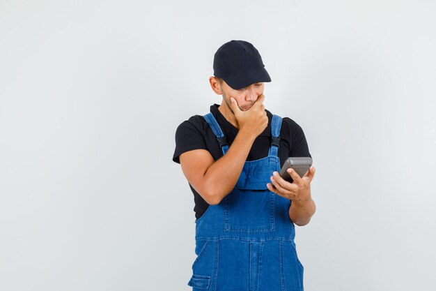 Jeune mécanicien en uniforme tenant la calculatrice avec la main sur la bouche et regardant pensif, vue de face.