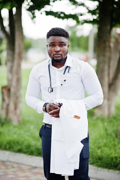 Jeune médecin afro-américain tenant un manteau blanc à portée de main avec un stéthoscope posé en plein air