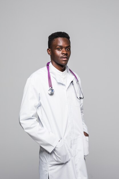 Jeune médecin afro-américain en uniforme blanc isolé sur fond blanc, debout avec les bras en poche à la recherche de professionnels et hautement compétent dans le domaine de la spécialisation médicale