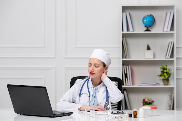 Jeune médecin blonde en blouse d'hôpital blanche et chapeau avec ordinateur fatigué tenant le cou