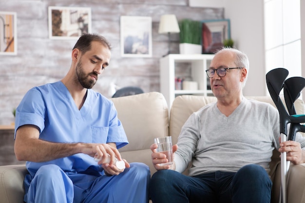 Jeune médecin caucasien dans une maison de retraite donnant des pilules à un homme âgé avec des béquilles. .