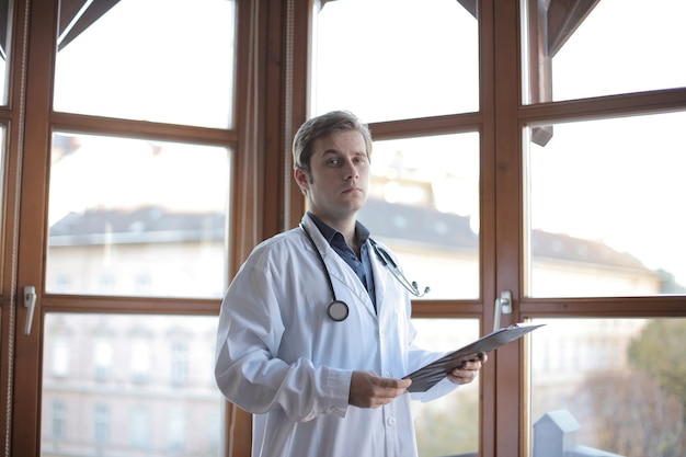 Jeune médecin debout dans son bureau avec des fenêtres sur l'arrière-plan