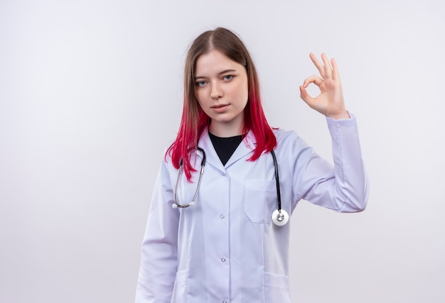 Jeune médecin femme portant une robe médicale stéthoscope montrant le geste okey sur mur blanc isolé