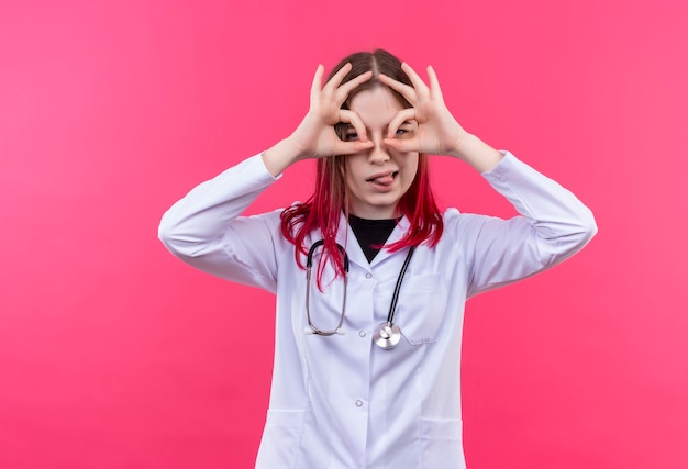 Jeune médecin femme portant une robe médicale stéthoscope montrant la langue et regarder le geste sur le mur isolé rose