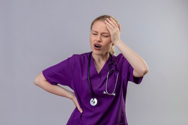 Jeune médecin malade portant une blouse médicale violette et un stéthoscope a mis sa main sur le front douloureux sur un mur blanc isolé