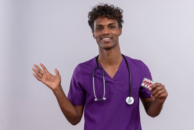 Un jeune médecin à la peau foncée beau sourire aux cheveux bouclés portant l'uniforme violet avec stéthoscope montrant des pilules