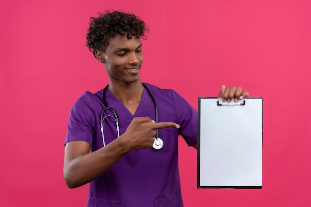 Photo gratuite un jeune médecin à la peau sombre beau sourire aux cheveux bouclés portant l'uniforme violet avec stéthoscope pointant avec l'index au presse-papiers avec une feuille de papier vierge