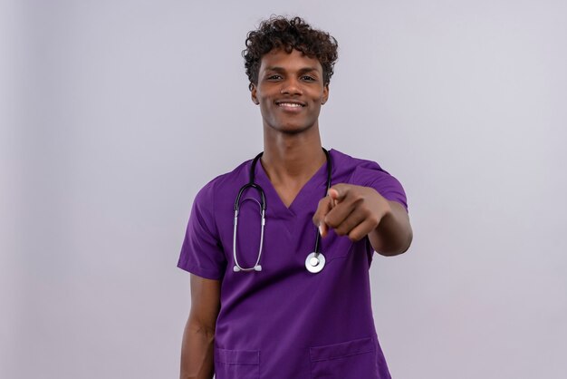 Un jeune médecin à la peau sombre beau sourire aux cheveux bouclés portant l'uniforme violet avec stéthoscope pointant vers la caméra avec l'index