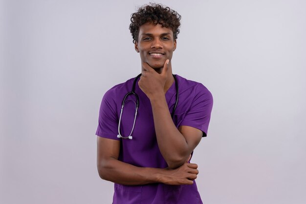 Un jeune médecin à la peau sombre beau sourire aux cheveux bouclés portant l'uniforme violet avec stéthoscope tenant la main sur le menton tout en