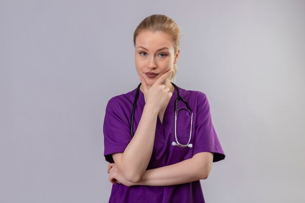 Jeune médecin portant une robe médicale violette et un stéthoscope a attrapé le menton sur un mur blanc isolé