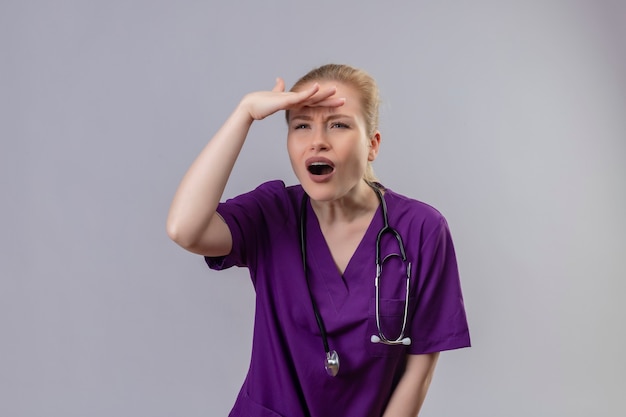 Jeune médecin portant une robe médicale violette et un stéthoscope à la distance avec la main sur un mur blanc isolé