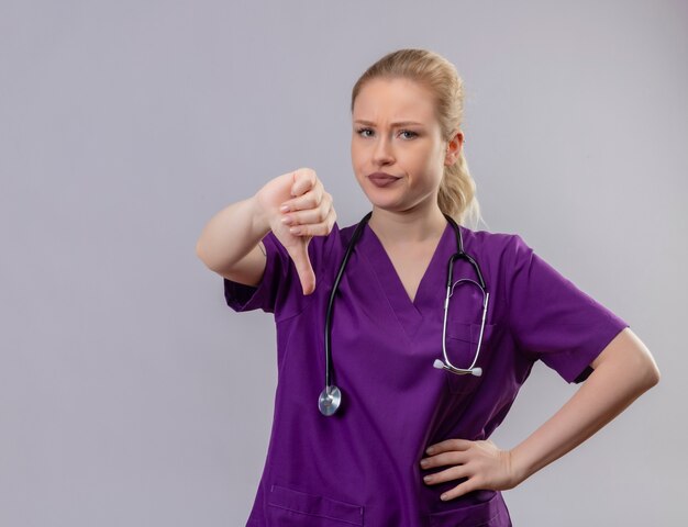 Jeune médecin portant une robe médicale violette et un stéthoscope son pouce vers le bas sur un mur blanc isolé