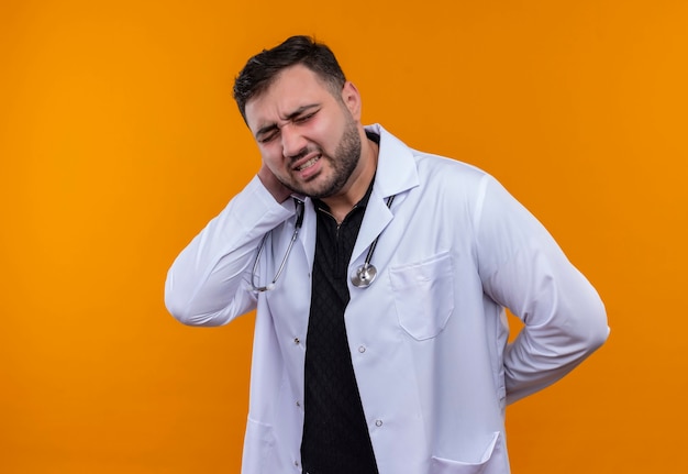 Jeune médecin de sexe masculin barbu portant une blouse blanche avec stéthoscope à la douleur au cou toucher mal