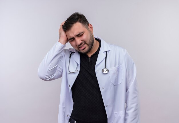 Jeune médecin de sexe masculin barbu portant une blouse blanche avec stéthoscope à la recherche d'un mal de tête fort touchant la tête