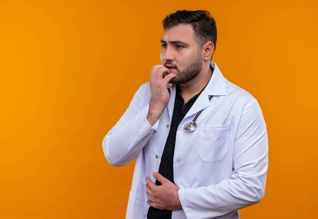 Jeune médecin de sexe masculin barbu portant une blouse blanche avec stéthoscope a souligné et nerveux mordant les ongles