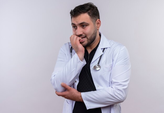 Jeune médecin de sexe masculin barbu portant une blouse blanche avec stéthoscope a souligné et nerveux de se ronger les ongles