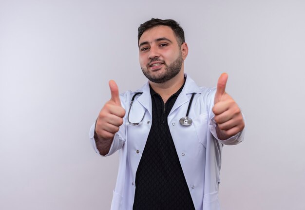 Jeune médecin de sexe masculin barbu portant une blouse blanche avec stéthoscope souriant montrant les pouces vers le haut avec les deux mains