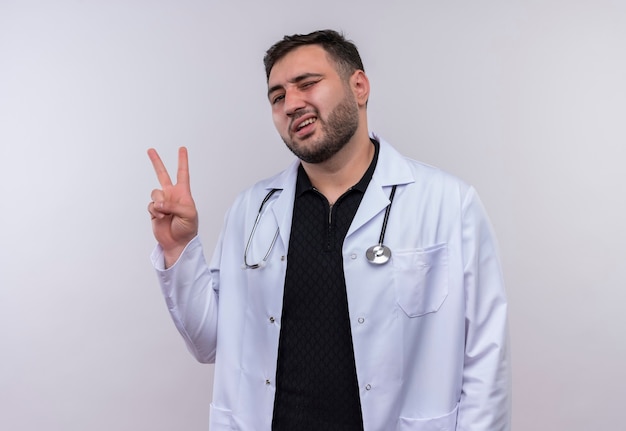 Jeune médecin de sexe masculin barbu portant un blouse blanche avec un stéthoscope en souriant et montrant le signe de la victoire
