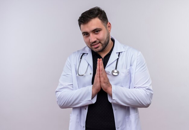 Jeune médecin de sexe masculin barbu portant une blouse blanche avec stéthoscope tenant les mains ensemble dans le geste de prière se sentir reconnaissant et heureux