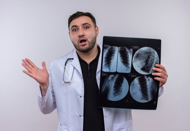 Jeune médecin de sexe masculin barbu portant un blouse blanche avec stéthoscope tenant des rayons x des poumons à la surprise et choqué