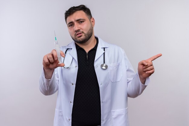 Jeune médecin de sexe masculin barbu portant une blouse blanche avec stéthoscope tenant la seringue à la confusion pointant avec le doigt sur le côté