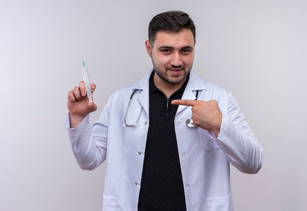 Jeune médecin de sexe masculin barbu portant une blouse blanche avec stéthoscope tenant la seringue pointant avec le doigt vers elle smiling confiant