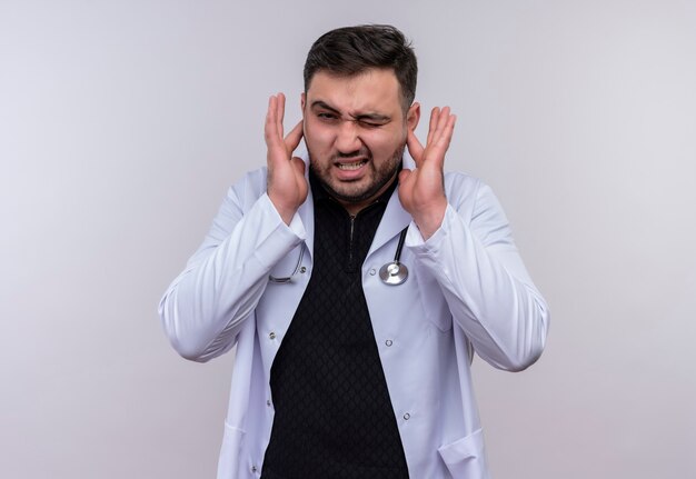 Jeune médecin de sexe masculin barbu portant un manteau blanc avec stéthoscope fermant les oreilles avec les doigts avec une expression agacée pour le bruit du son fort