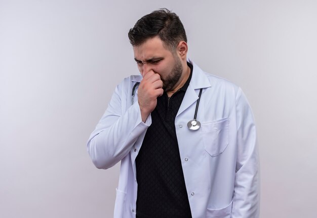 Jeune médecin de sexe masculin barbu portant un manteau blanc avec stéthoscope fermant son nez avec les doigts sentant un parfum désagréable