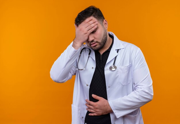 Jeune médecin de sexe masculin barbu portant un manteau blanc avec stéthoscope lookin malade se sentir malade toucher la tête