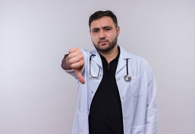 Jeune médecin de sexe masculin barbu portant un manteau blanc avec un stéthoscope à mécontent montrant l'aversion
