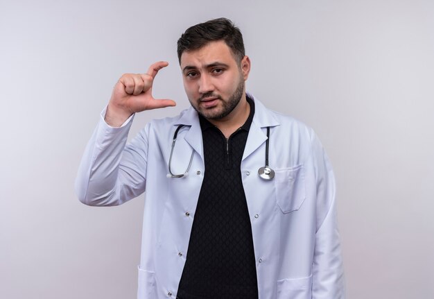 Jeune médecin de sexe masculin barbu portant un manteau blanc avec stéthoscope montrant le geste de taille avec la main, symbole de mesure