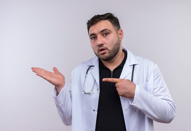 Jeune médecin de sexe masculin barbu portant un manteau blanc avec stéthoscope présent avec le bras oh sa main pointant avec l'index au bras