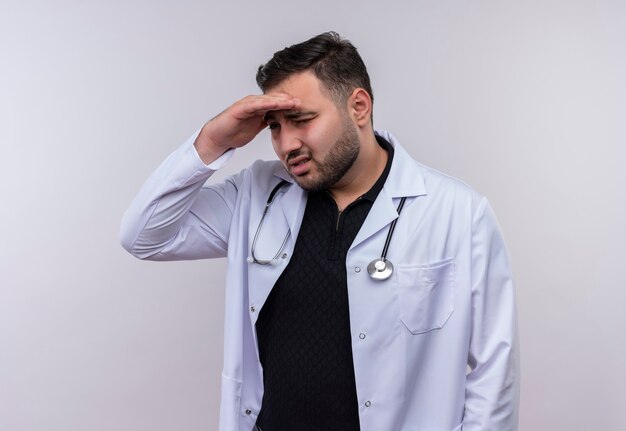 Jeune médecin de sexe masculin barbu portant un manteau blanc avec stéthoscope à la recherche de loin avec la main sur la tête pour regarder quelque chose ou quelqu'un