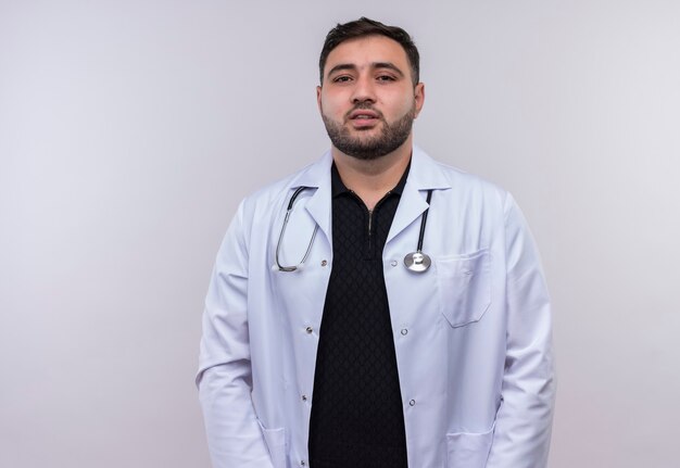 Jeune médecin de sexe masculin barbu portant un manteau blanc avec stéthoscope regardant la caméra avec une expression confiante
