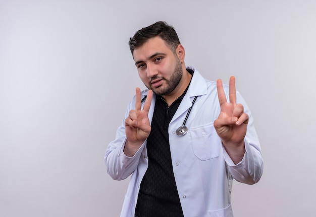 Jeune médecin de sexe masculin barbu portant un manteau blanc avec stéthoscope souriant confiant montrant des signes de victoire avec les deux mains