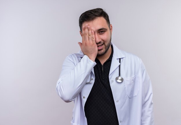 Jeune médecin de sexe masculin barbu portant un manteau blanc avec stéthoscope souriant couvrant un œil avec palm