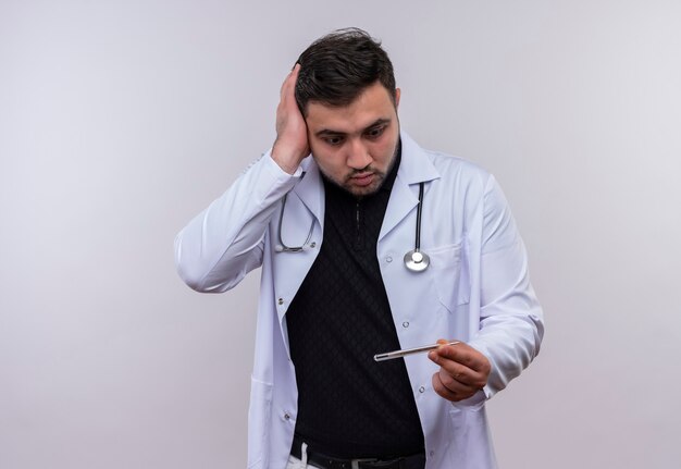 Jeune médecin de sexe masculin barbu portant un manteau blanc avec un stéthoscope tenant un thermomètre en le regardant très émouvant et inquiet