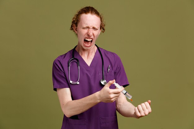 Jeune médecin de sexe masculin en colère portant l'uniforme avec stéthoscope tenant la seringue isolé sur fond vert
