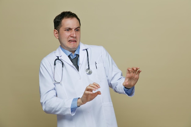 Jeune médecin de sexe masculin dégoûté portant une robe médicale et un stéthoscope autour du cou regardant le côté ne faisant aucun geste isolé sur fond vert olive