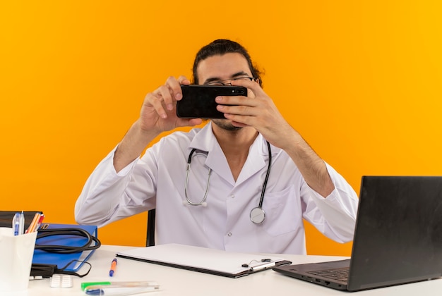 Jeune médecin de sexe masculin avec des lunettes médicales portant une robe médicale avec stéthoscope assis au bureau