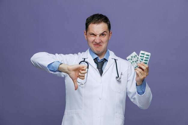 Jeune médecin de sexe masculin mécontent portant une robe médicale et un stéthoscope autour du cou regardant la caméra montrant des paquets de pilules et le pouce vers le bas isolé sur fond violet